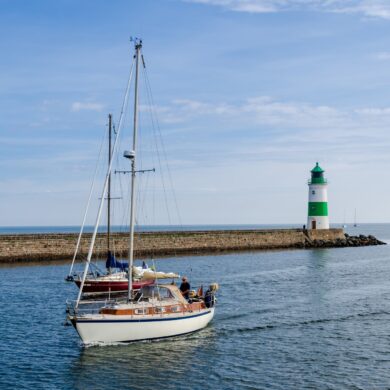 Zwei Segelboote fahren vor dem grün-weißen Leuchtturm in Kappeln an der Ostseeküste übers Wasser.