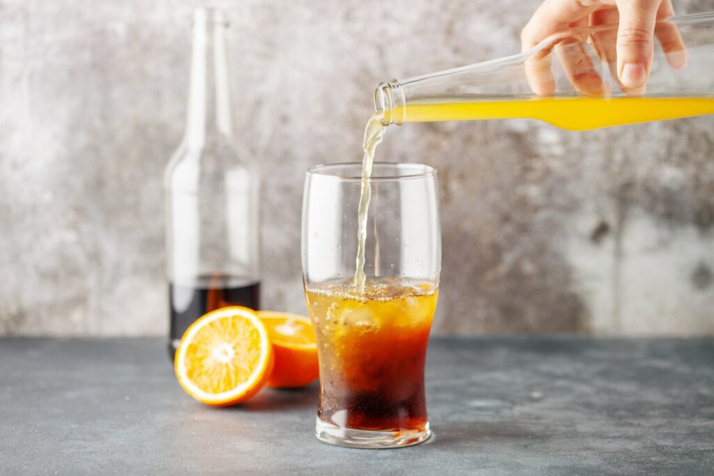 A hand pours orange soda into a glass with cola