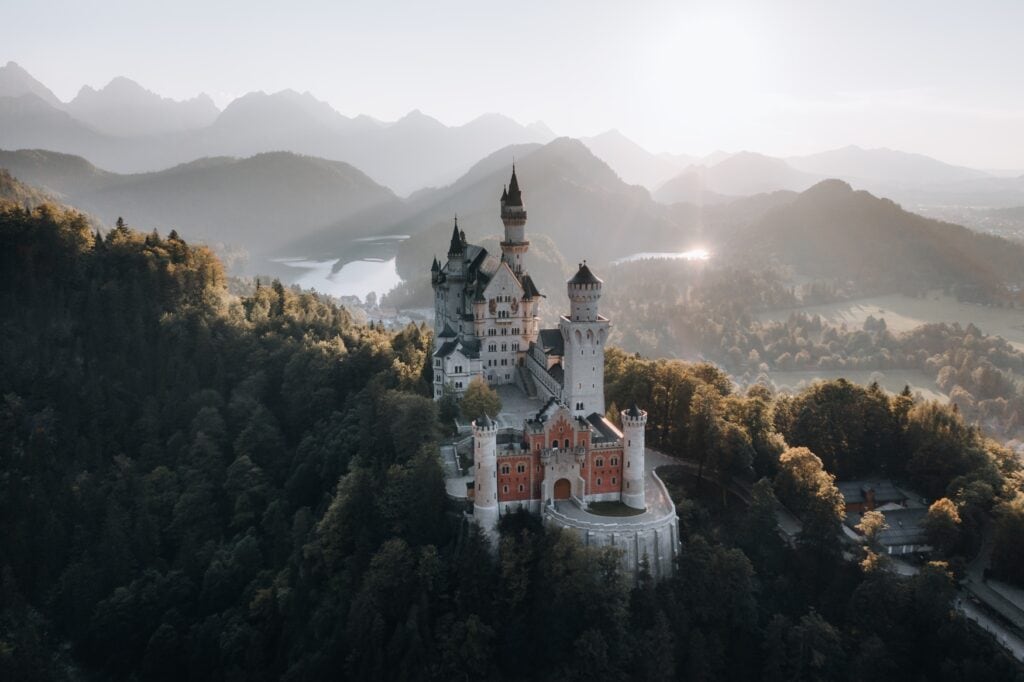 Schloss Neuschwanstein in einer Drohnenaufnahme. Hinter dem Schloss liegt ein Tal im Morgennebel.