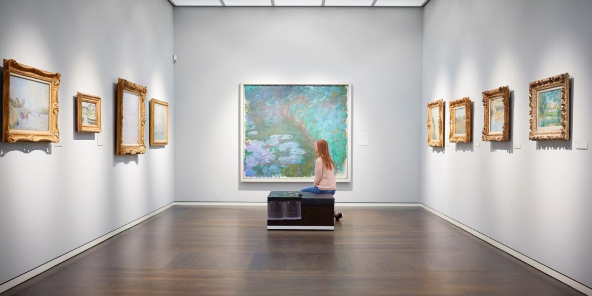 Eine Frau sitzt auf einer Bank in einem weißen Ausstellungssaal mit mehreren Bildern an der Wand und schaut auf das große Aquarellgemälde genau vor ihr.