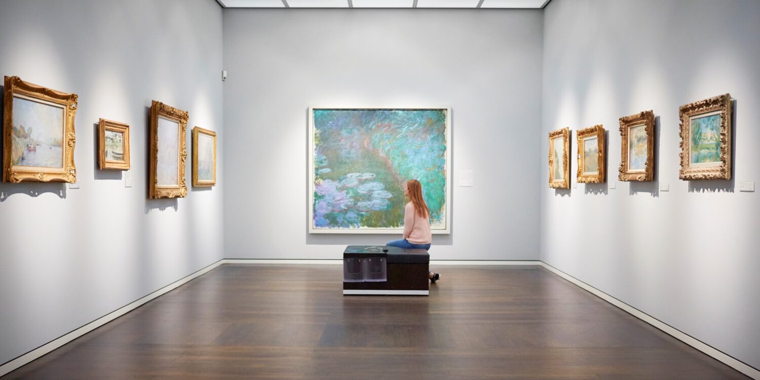 Eine Frau sitzt auf einer Bank in einem weißen Ausstellungssaal mit mehreren Bildern an der Wand und schaut auf das große Aquarellgemälde genau vor ihr.