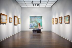 Eine Frau sitzt auf einer Bank in einem weißen Ausstellungssaal mit mehreren Bildern an der Wand und schaut auf das große Aquarellgemälde genau vor ihr.