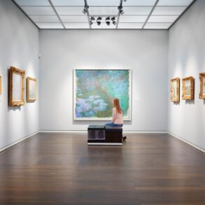 Eine Frau sitzt auf einer Bank in einem weißen Ausstellungssaal mit mehreren Bildern an der Wand und schaut auf das große Aquarellgemälde genau vor ihr.