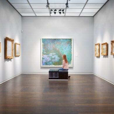 Eine Frau sitzt auf einer Bank in einem weißen Ausstellungssaal mit mehreren Bildern an der Wand und schaut auf das große Aquarellgemälde genau vor ihr.