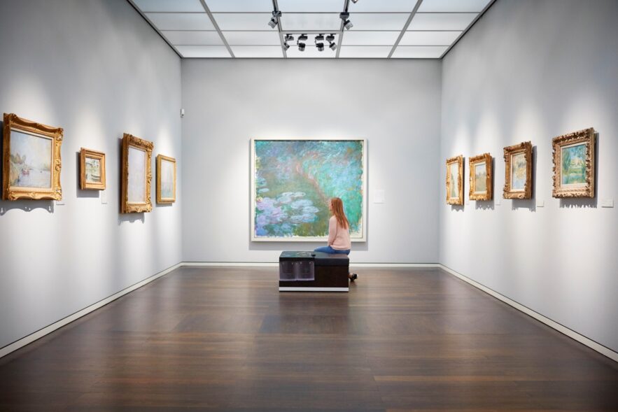 Eine Frau sitzt auf einer Bank in einem weißen Ausstellungssaal mit mehreren Bildern an der Wand und schaut auf das große Aquarellgemälde genau vor ihr.