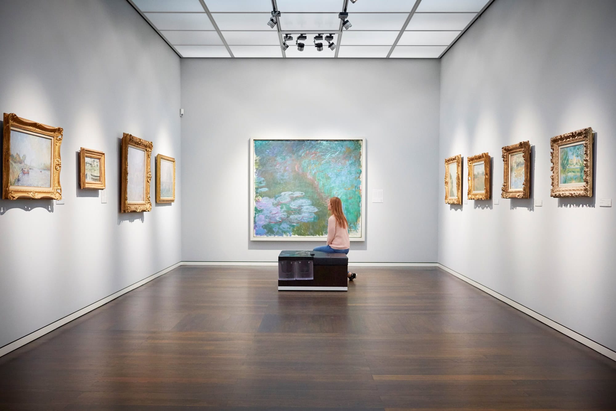 Eine Frau sitzt auf einer Bank in einem weißen Ausstellungssaal mit mehreren Bildern an der Wand und schaut auf das große Aquarellgemälde genau vor ihr.