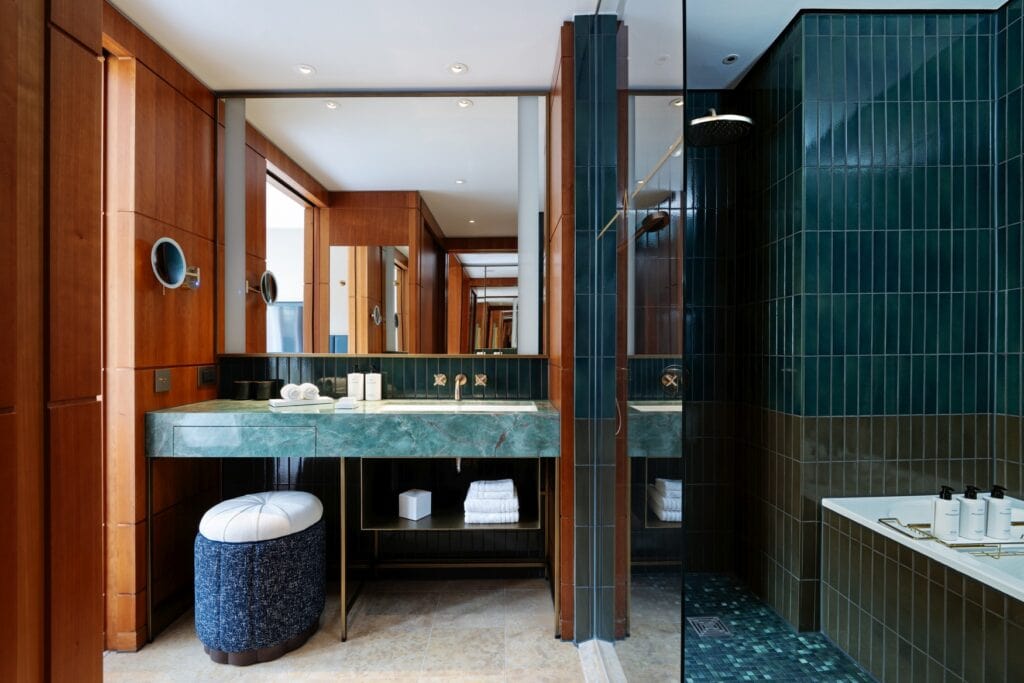 Ein Bad in einem Hotelzimmer. Auf der linken Seite ein Waschtisch aus grünem Marmor, darüber ein großer, rechteckiger Spiegel. Auf der rechten Seite der Badbereich mit Dusche und Badewanne abgetrennt durch eine Glasscheibe. Der Badbereich ist mit grünen Fliesen ausgekleidet.