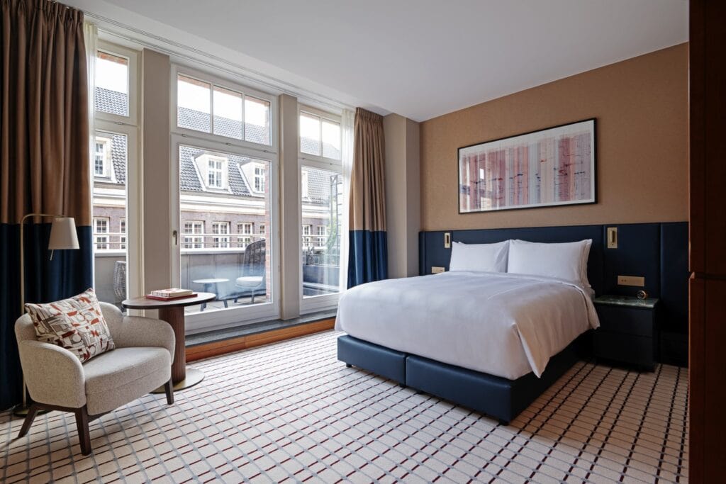 Ein Hotelzimmer, zur rechten ein blaues Doppelbett mit weißen Bezügen, dahinter eine hellbraune Wand mit einem Gemälde. Rechts ein grauer Polstersessel mit Beistelltisch. Die drei großen Fenster führen auf einen Balkon. Der Boden ist mit einem weißen Teppich mit blauen Details ausgelegt.