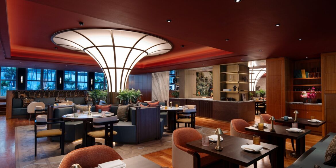 Ein modern eingerichtetes Restaurant mit vielen Sitzgruppen. Die Polsterstühle in der Reihe im Vordergrund sind orange, dahinter stehen auch cremefarbene. Die Decke ist rot, wodurch eine gedeckte Stimmung entsteht. Mitten im Restaurant steht eine nach oben breiter werdende, runde Lichtsäule, deren Panele nur von schmalen schwarzen Balken getrennt sind.