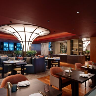 Ein modern eingerichtetes Restaurant mit vielen Sitzgruppen. Die Polsterstühle in der Reihe im Vordergrund sind orange, dahinter stehen auch cremefarbene. Die Decke ist rot, wodurch eine gedeckte Stimmung entsteht. Mitten im Restaurant steht eine nach oben breiter werdende, runde Lichtsäule, deren Panele nur von schmalen schwarzen Balken getrennt sind.