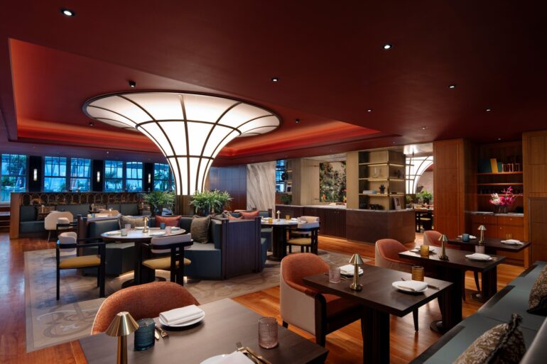 Ein modern eingerichtetes Restaurant mit vielen Sitzgruppen. Die Polsterstühle in der Reihe im Vordergrund sind orange, dahinter stehen auch cremefarbene. Die Decke ist rot, wodurch eine gedeckte Stimmung entsteht. Mitten im Restaurant steht eine nach oben breiter werdende, runde Lichtsäule, deren Panele nur von schmalen schwarzen Balken getrennt sind.