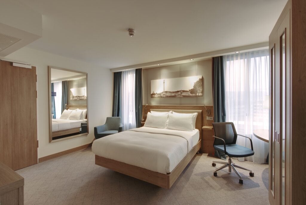 Ein Hotelzimmer mit Teppichboden, einem "schwebenden" Doppelbett in der Mitte, Holzkopfteil, einem Schrank zur rechten, der Tür zur linken und zwei Fenstern hinter dem Bett.