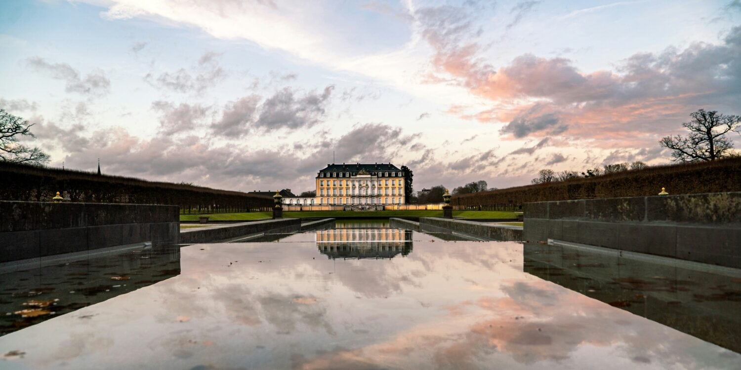 Hinter einem langen, flachen Schlossteich steht in der Ferne die gelbe Fassade des Schlosses Augustusburg in Brühl, das sich im Wasser spiegelt. Darüber geht die Sonne an einem leicht bewölkten Himmel unter.