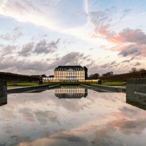 Hinter einem langen, flachen Schlossteich steht in der Ferne die gelbe Fassade des Schlosses Augustusburg in Brühl, das sich im Wasser spiegelt. Darüber geht die Sonne an einem leicht bewölkten Himmel unter.