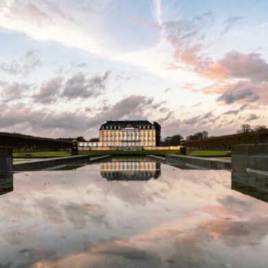 Hinter einem langen, flachen Schlossteich steht in der Ferne die gelbe Fassade des Schlosses Augustusburg in Brühl, das sich im Wasser spiegelt. Darüber geht die Sonne an einem leicht bewölkten Himmel unter.