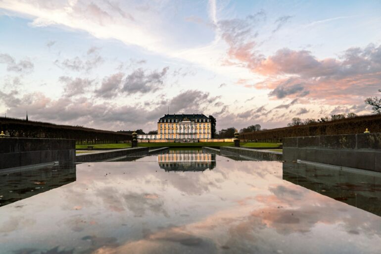 Hinter einem langen, flachen Schlossteich steht in der Ferne die gelbe Fassade des Schlosses Augustusburg in Brühl, das sich im Wasser spiegelt. Darüber geht die Sonne an einem leicht bewölkten Himmel unter.