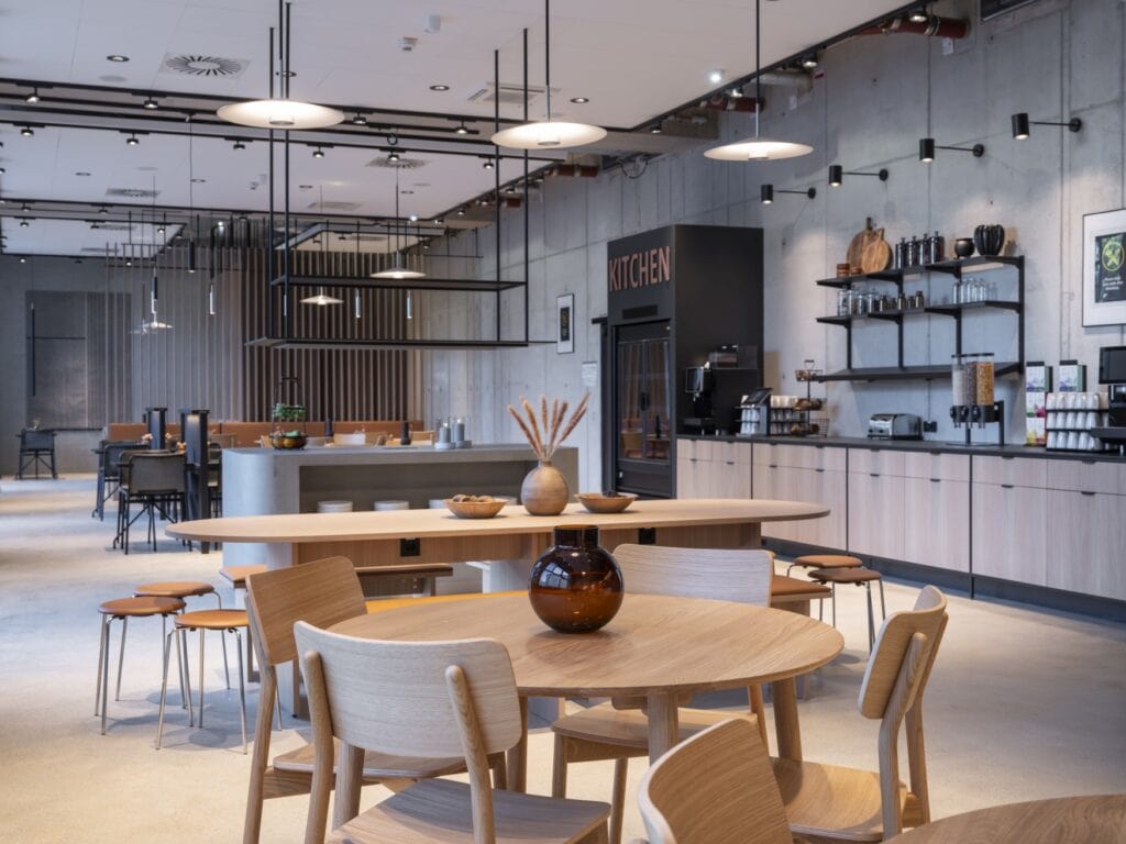 Eine Hotellounge, eingerichtet wie ein moderner Speisesaal. Mehrere Sitzgruppen mit Holzmöbeln stehen in einem grauen Raum, darüber Metalllampen. Zur rechten eine lange Anrichte mit einer Kaffee- und Teestation sowie Müslispendern und einem Kühlschrank. Alles ist in einem kühlen, minimalistischen Design gehalten.