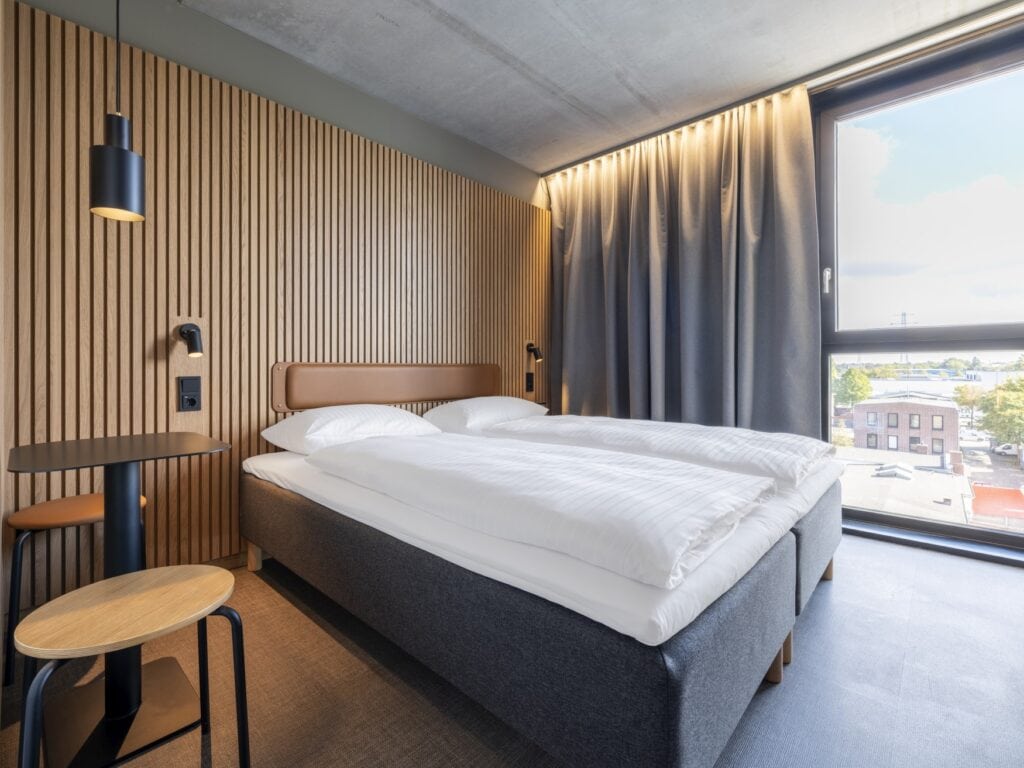 Ein graues Doppelbett mit weißen Bezügen in einem Hotelzimmer. Die Wand hinter dem Kopfteil ist mit hellen Holzlamellen verkleidet, zur linken des Betts ein einfacher Metalltisch mit zwei Holzhockern. Zur rechten des Bettes ein großes Fenster mit Ausblick.
