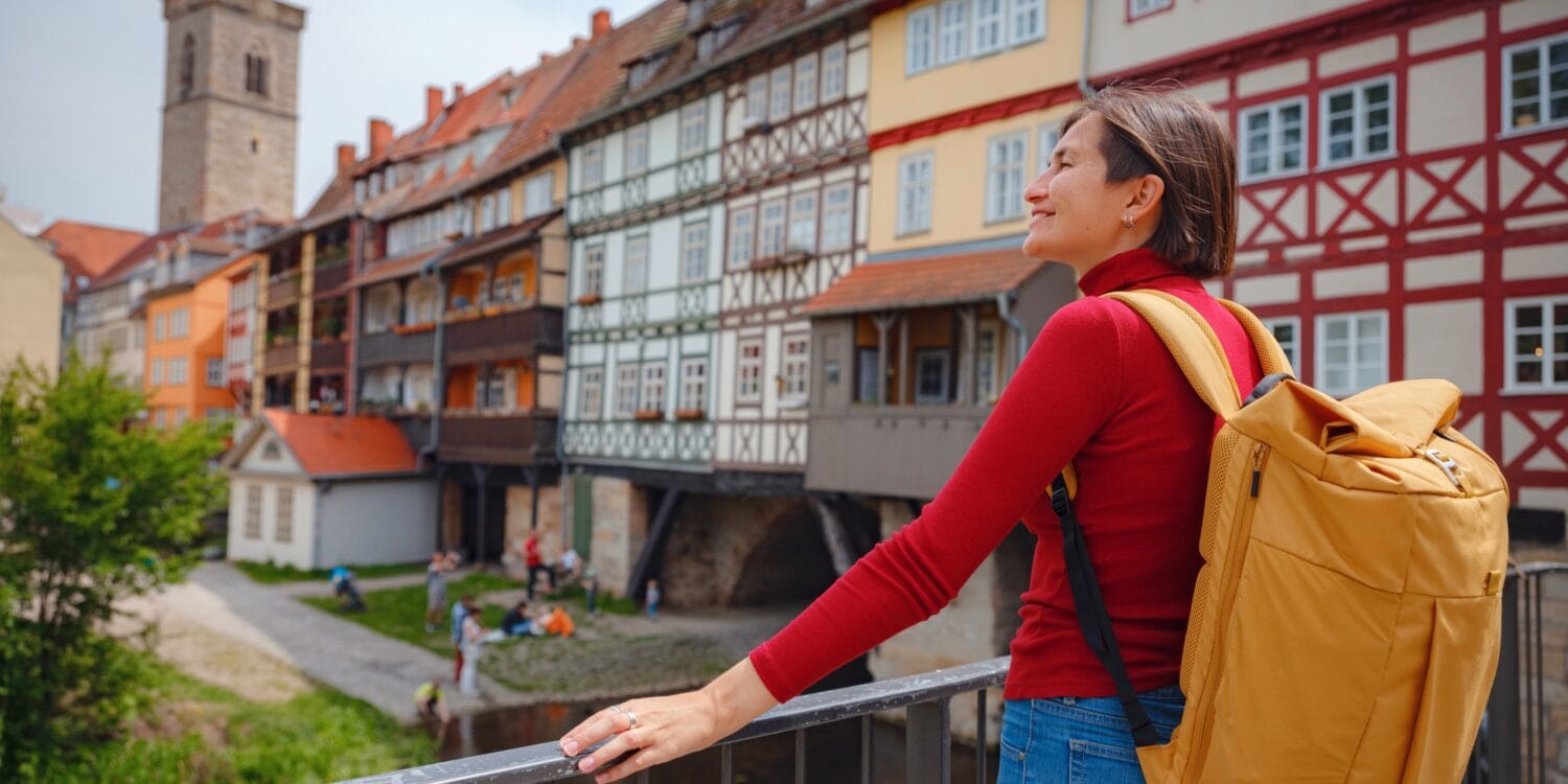 Eine Frau mit Rucksack steht an einem Geländer und blickt nach links aus dem Bild; rechts von ihr ziehen sich die bunten Fachwerkfassaden der Krämerbrücke, einer der Sehenswürdigkeiten in Erfurt, in den Hintergrund des Bilds