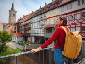 Eine Frau mit Rucksack steht an einem Geländer und blickt nach links aus dem Bild; rechts von ihr ziehen sich die bunten Fachwerkfassaden der Krämerbrücke, einer der Sehenswürdigkeiten in Erfurt, in den Hintergrund des Bilds