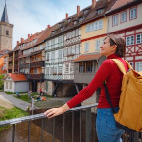 Eine Frau mit Rucksack steht an einem Geländer und blickt nach links aus dem Bild; rechts von ihr ziehen sich die bunten Fachwerkfassaden der Krämerbrücke, einer der Sehenswürdigkeiten in Erfurt, in den Hintergrund des Bilds