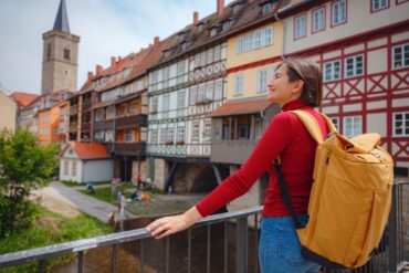Eine Frau mit Rucksack steht an einem Geländer und blickt nach links aus dem Bild; rechts von ihr ziehen sich die bunten Fachwerkfassaden der Krämerbrücke, einer der Sehenswürdigkeiten in Erfurt, in den Hintergrund des Bilds