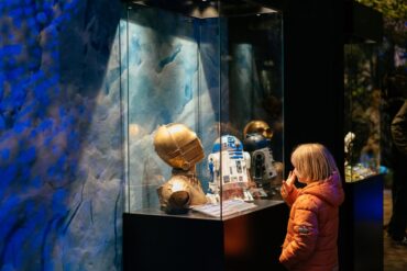 A child stands in front of a display case containing the head of C3PO at an exhibition.