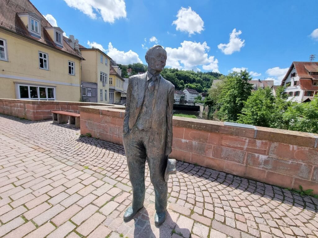 Eine lebensgroße Bronzestatue des Schriftstellers Hermann Hesse steht vor einem kleinen Mäuerchen, dahinter einige Bäume