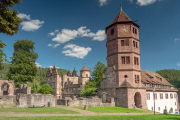 Eine mittelalterliche Kloster- und Schlossanlage aus rotem Sandstein: Links einige verfallene Mauern und der Rest eines Krezgangs, mittig ein vierstöckiger Palas ohne Dach, rechts ein hoher achteckiger Turm mit angebrachter Uhr