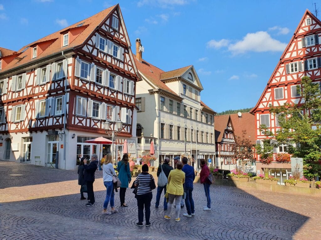 Eine Gruppe Menschen im sommerlichen Calw steht auf einem Platz und schaut auf eine Reihe von der Sonne angeschienener, sehr gut erhaltener Fachwerkhäuser
