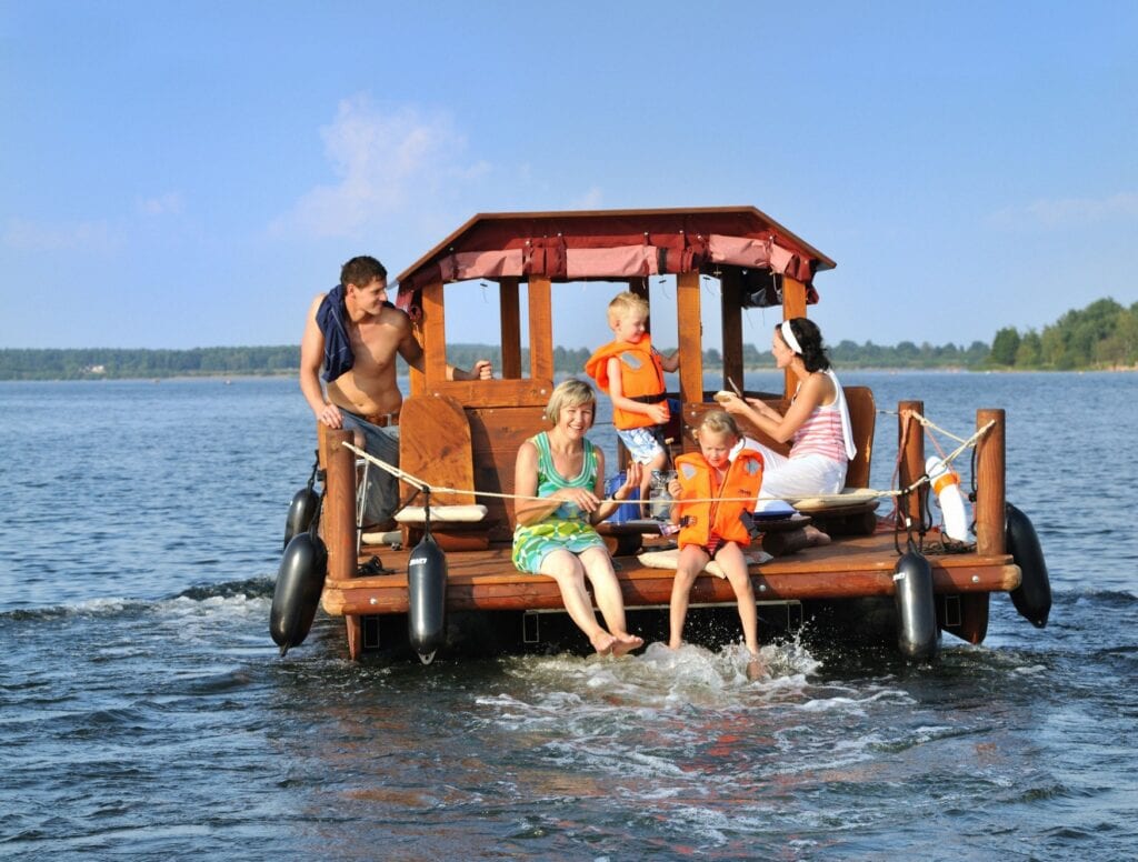 Drei Erwachsene und zwei Kinder stehen und sitzen auf einem hölzernen Floß mit Verdeck und fahren über einen See. Die Kinder tragen Schwimmwesten, die Erwachsenen Badekleidung. Eine Frau reicht einem der Kinder ein Butterbrot, eine andere Frau und ein Kind lassen ihre Füße in die Fahrtwellen hängen. Ein rund um das Floß gespanntes Tau sorgt dafür, dass alle jederzeit Wissen, wo das Floß endet.