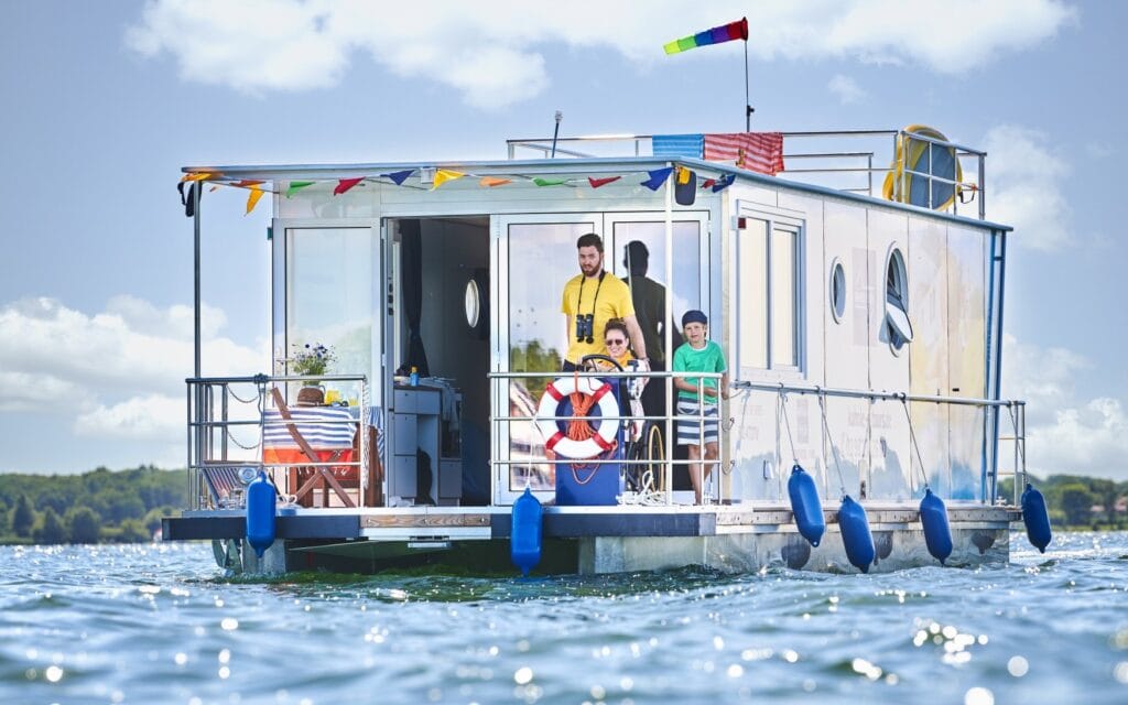 Ein weißes Hausboot, das mit bunten Wimpeln dekoriert ist, fährt auf einem See. An Deck stehen ein Mann in einem gelben T-Shirt und ein Kind. Am Steuer sitzt eine Frau im Rollsthul mit Sonnebrille, die das Hausboot fährt. Rund um das Boot glitzert die Sonne auf der Wasseroberfläche, im Hintergrund sieht man ein bewaldetes Ufer
