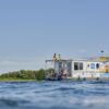 Ein weißes Hausboot, das mit bunten Wimpeln dekoriert ist, fährt auf einem See. Auf dem Oberdeck sitzen ein Mann in einem gelben T-Shirt und ein Kind. Am Steuer sitzt eine Frau im Rollstuhl mit Sonnebrille, die das Hausboot fährt. Rund um das Boot glitzert die Sonne auf der Wasseroberfläche, im Hintergrund sieht man ein bewaldetes Ufer