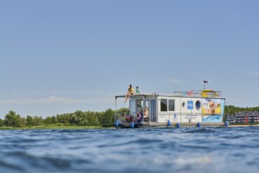 Ein weißes Hausboot, das mit bunten Wimpeln dekoriert ist, fährt auf einem See. Auf dem Oberdeck sitzen ein Mann in einem gelben T-Shirt und ein Kind. Am Steuer sitzt eine Frau im Rollstuhl mit Sonnebrille, die das Hausboot fährt. Rund um das Boot glitzert die Sonne auf der Wasseroberfläche, im Hintergrund sieht man ein bewaldetes Ufer