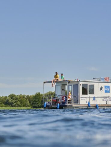 Ein weißes Hausboot, das mit bunten Wimpeln dekoriert ist, fährt auf einem See. Auf dem Oberdeck sitzen ein Mann in einem gelben T-Shirt und ein Kind. Am Steuer sitzt eine Frau im Rollstuhl mit Sonnebrille, die das Hausboot fährt. Rund um das Boot glitzert die Sonne auf der Wasseroberfläche, im Hintergrund sieht man ein bewaldetes Ufer