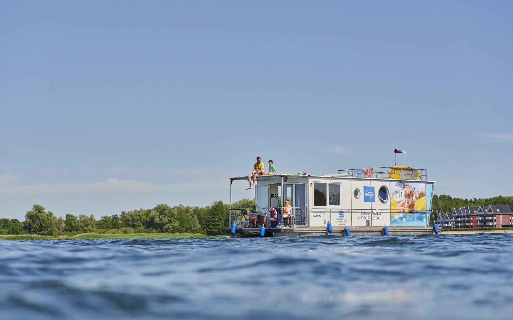 Ein weißes Hausboot, das mit bunten Wimpeln dekoriert ist, fährt auf einem See. Auf dem Oberdeck sitzen ein Mann in einem gelben T-Shirt und ein Kind. Am Steuer sitzt eine Frau im Rollstuhl mit Sonnebrille, die das Hausboot fährt. Rund um das Boot glitzert die Sonne auf der Wasseroberfläche, im Hintergrund sieht man ein bewaldetes Ufer