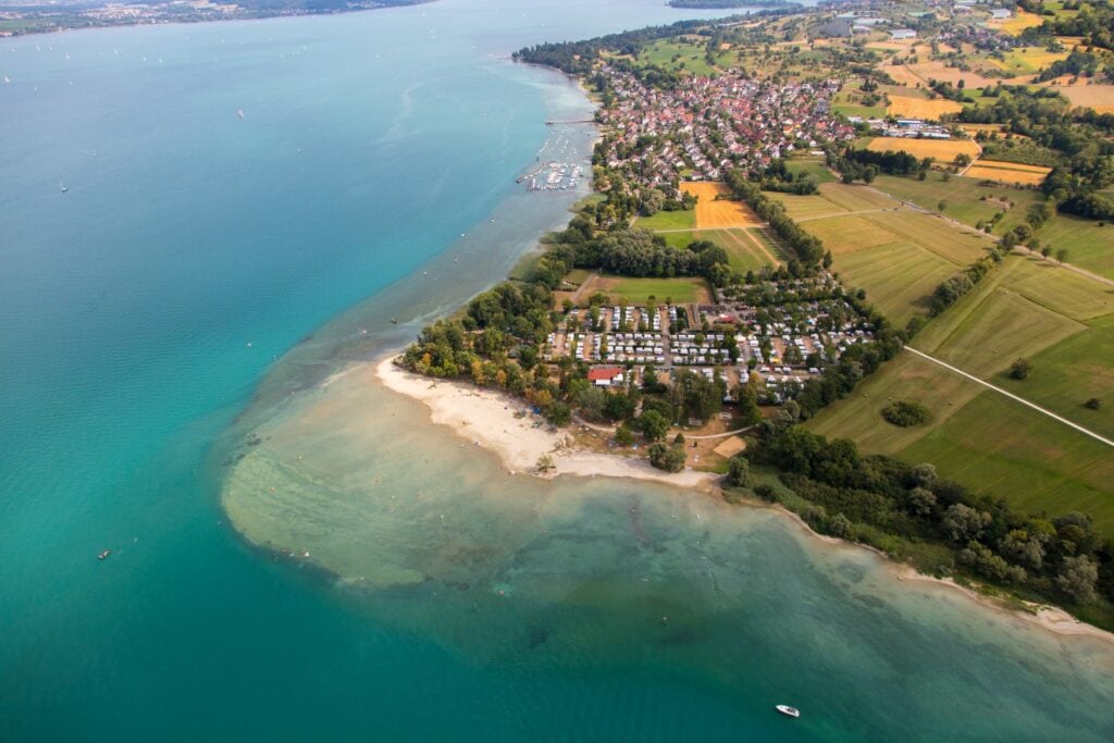 Luftaufnahme des Bodensees links und der Uferseite rechts; am Ufer ist ein heller Sandstrand im Fokus, dahinter Ortschaften und Felder