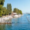 Ein Strandbad; links führen Treppen in den Rheinarm bei Konstanz, viele Leute in Badekleidung; im Hintergrund führt eine Brücke über den Fluss