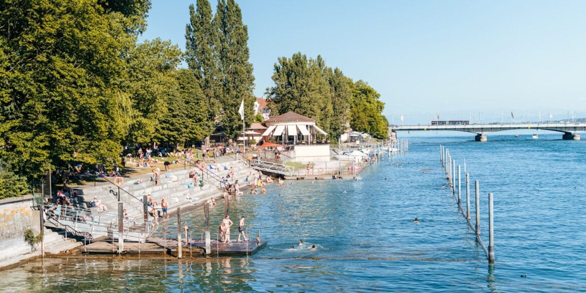 Ein Strandbad; links führen Treppen in den Rheinarm bei Konstanz, viele Leute in Badekleidung; im Hintergrund führt eine Brücke über den Fluss