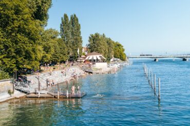 Ein Strandbad; links führen Treppen in den Rheinarm bei Konstanz, viele Leute in Badekleidung; im Hintergrund führt eine Brücke über den Fluss