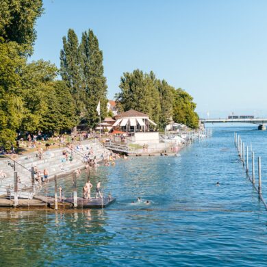Ein Strandbad; links führen Treppen in den Rheinarm bei Konstanz, viele Leute in Badekleidung; im Hintergrund führt eine Brücke über den Fluss