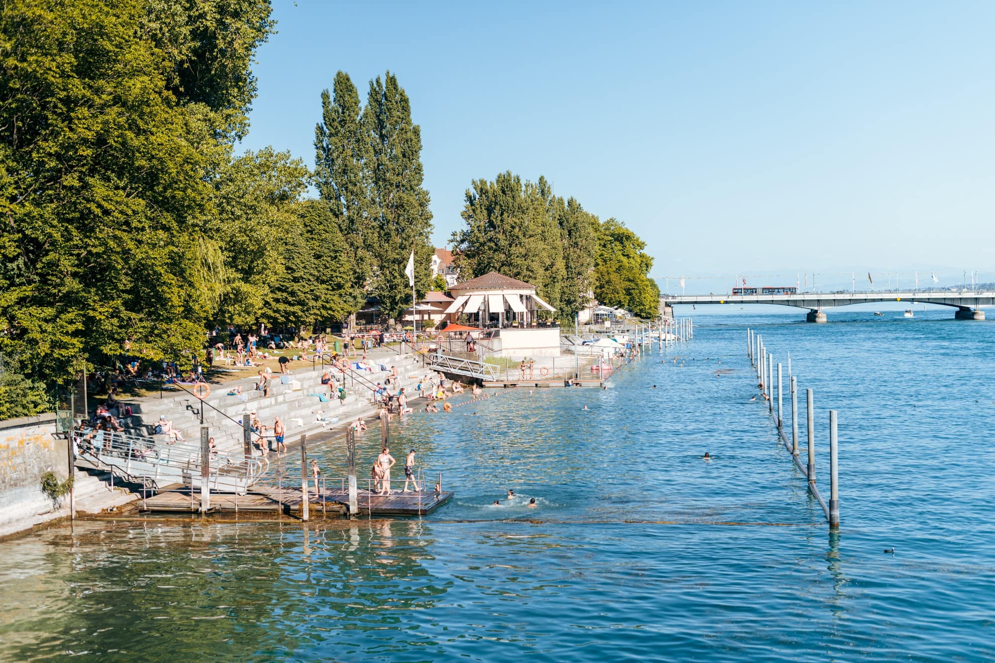 Ein Strandbad; links führen Treppen in den Rheinarm bei Konstanz, viele Leute in Badekleidung; im Hintergrund führt eine Brücke über den Fluss