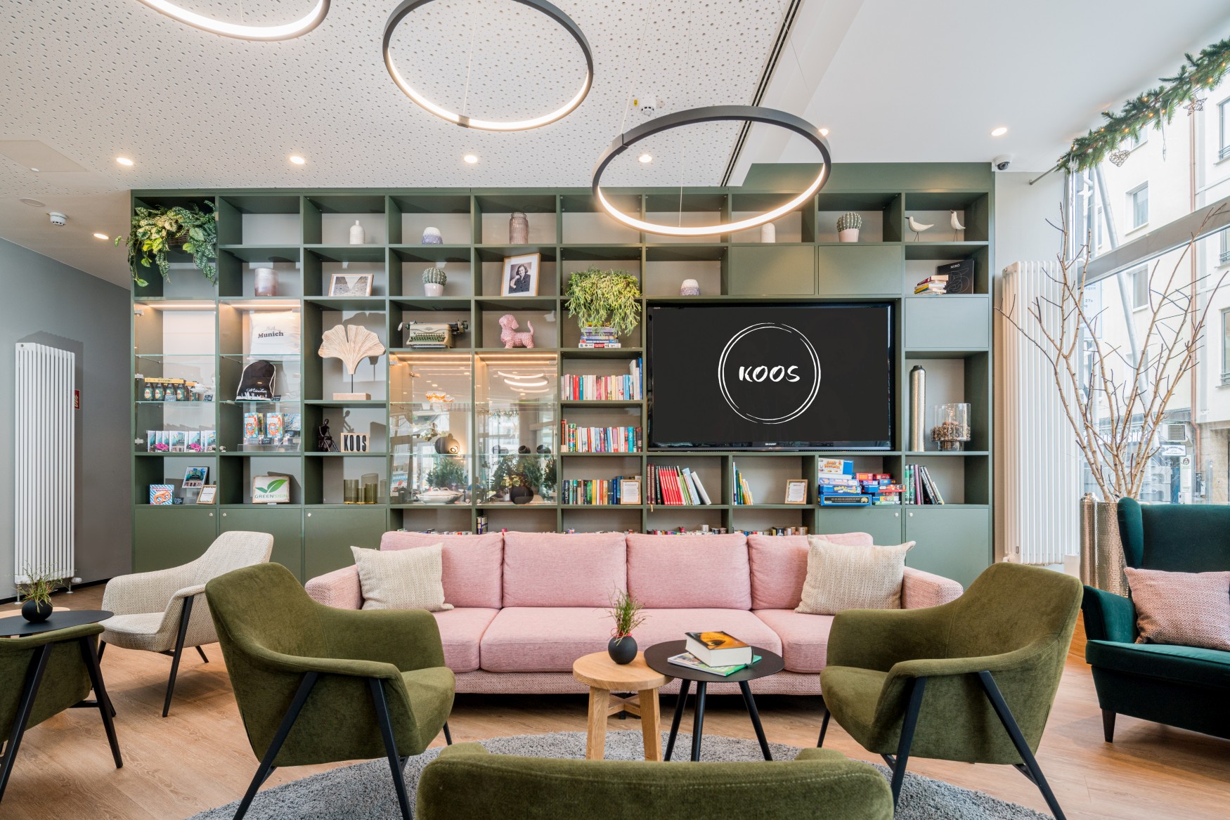 A hotel lobby furnished as a living room; in the foreground, a set of green armchairs and a pink sofa; in the background, a very tall bookcase with books and decorative objects. Integrated into it is a large television showing the “KOOS” logo.