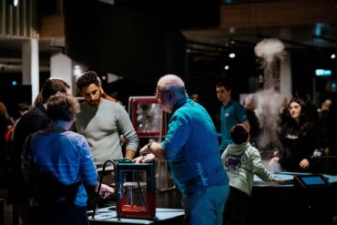 Mehrere Personen stehen in einem nächtlichen Museum um angestrahlte Technik-Exponate herum