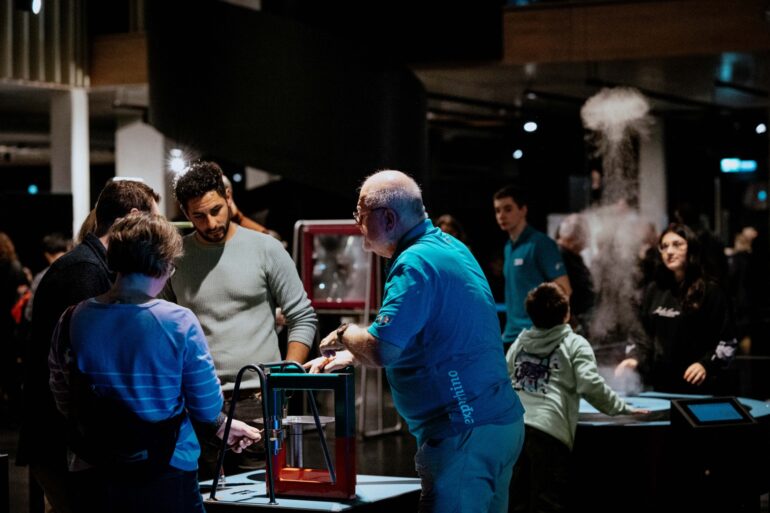 Mehrere Personen stehen in einem nächtlichen Museum um angestrahlte Technik-Exponate herum