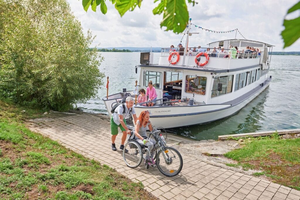 Eine Person in einem Rollstuhl mit Handbike-Erweiterung wird von einer zweiten Person von einem Fährboot mit der Aufschrift "MS Altmühlsee", das am Ufer eines Sees ankert, weggeschoben. Dahinter folgt ein weiteres Paar mit Rollstuhl, das gerade das Schiff verlässt. Auf dem Oberdeck sitzen noch viele Gäste.