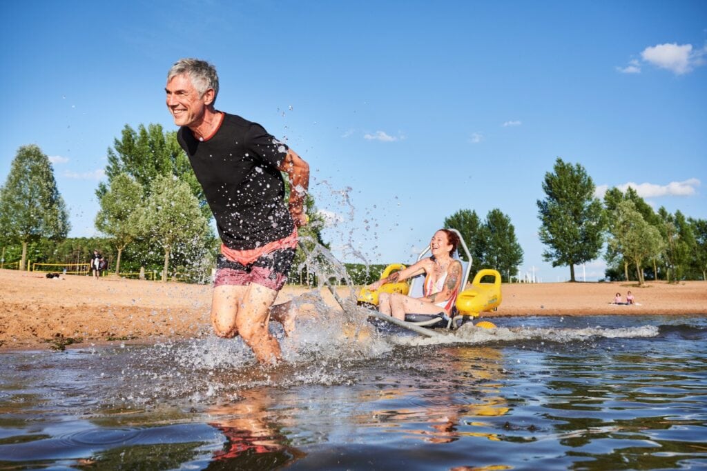 Ein Mann zieht eine Frau in einem Strandrollstuhl schwungvoll durch das ufernahe Wasser eines Sees, wobei viel Wasser aufspritzt. Beide haben sichtlich Spaß. Im Hintergrund erstreckt sich ein Sandstrand mit einigen Badegästen, dahinter mehrere grüne Bäume