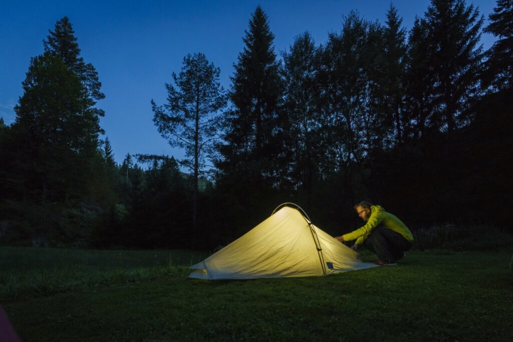 A man enters a tent lit from inside in a forest at night.