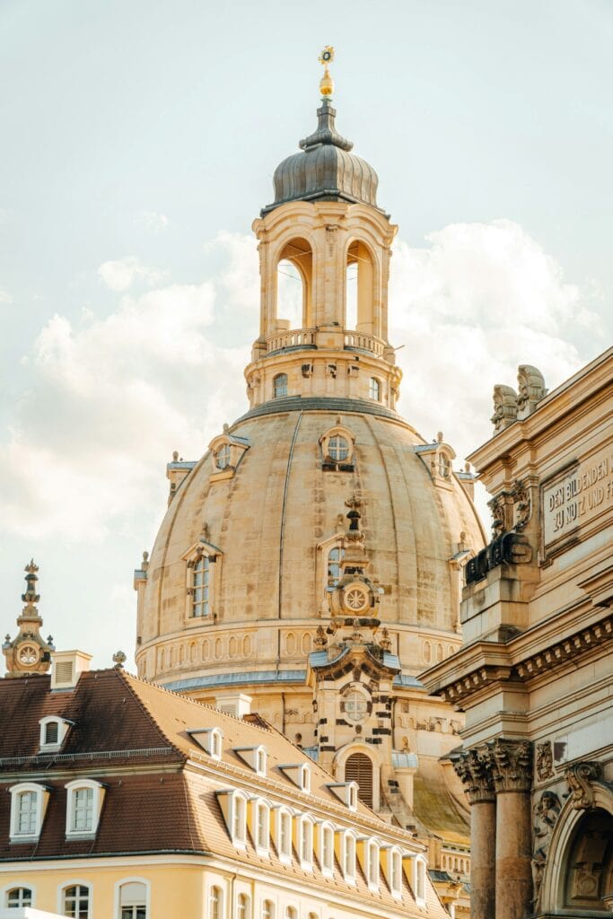 Zoom-Aufnahme der hellen Kuppel der Dresdner Frauenkirche vor blauem Himmel. Links davor das Dach eines barocken Wohngebäudes, rechts im Vordergrund ragt ein Triumphbogen, das Kronentor des Dresdner Zwingers, ins Bild.