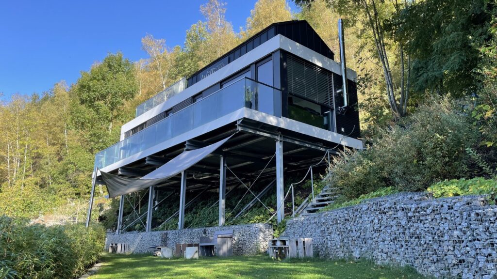 Ein von Metallpfeiler gestütztes Haus an einem bewaldeten Hang; bodentiefe Glasfenster, davor ein Balkon mit Glasgeländer; auf dem Metalldach ist der Schriftzug "Williadukt" angebracht